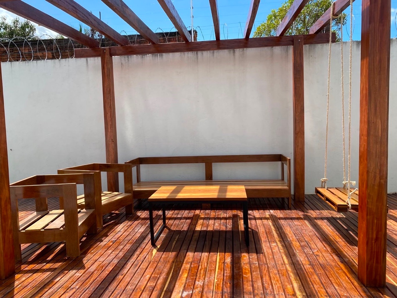 Deck externo com pergolado de madeira, mesa central, sofás e balanço